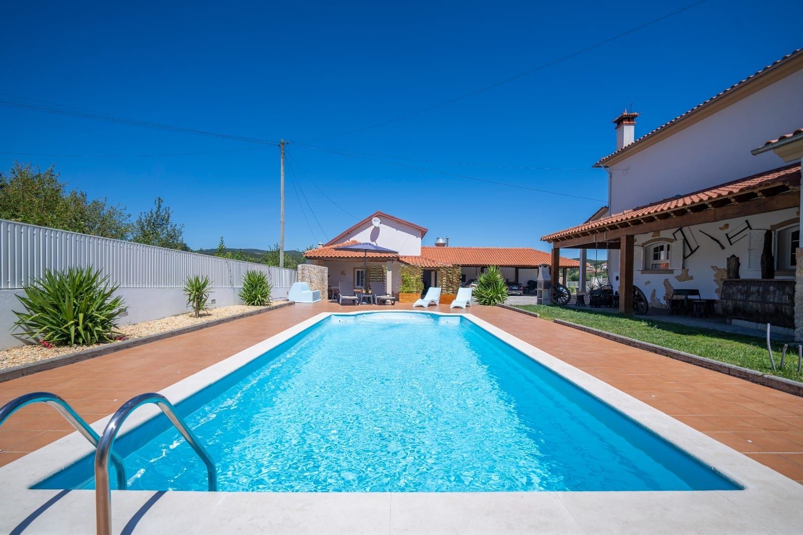 Casa de férias em Mortágua com piscina azul cristalina, rodeada por um jardim bem cuidado, área de lazer com bar e espreguiçadeiras. A arquitetura da casa é tradicional, com um telhado de telhas vermelhas e paredes brancas.